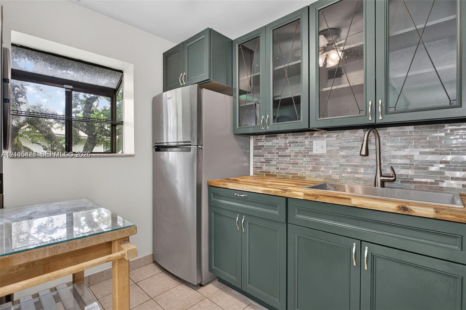 Kitchen featuring blue-green shaker cabinetry with glass-front uppers, butcher block countertops, stainless steel undermount sink with gooseneck faucet, and mosaic tile backsplash