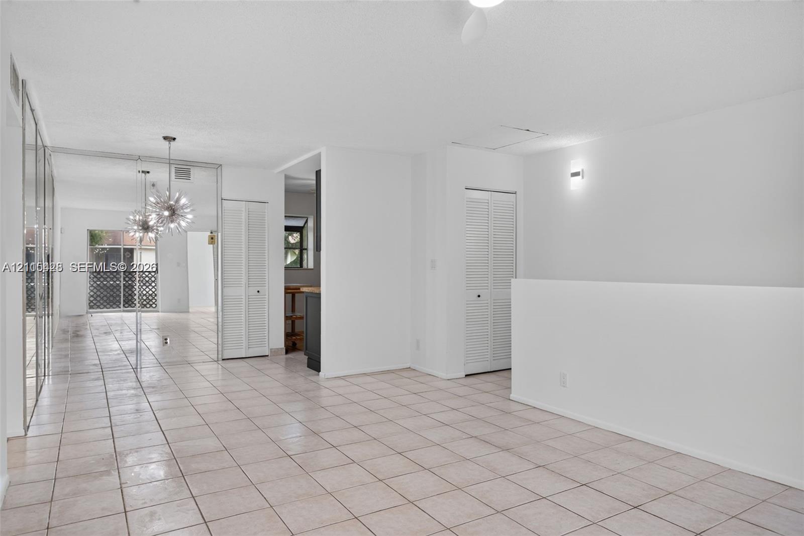Open-concept interior featuring light-toned tile flooring, floor-to-ceiling mirrored wall panels, louvered-door closets, and a decorative multi-bulb pendant fixture