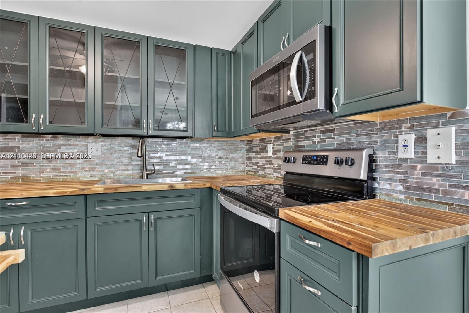 Kitchen featuring custom-color cabinetry with glass-front uppers, wood-finish countertops, stainless steel appliances, and multi-tone horizontal tile backsplash