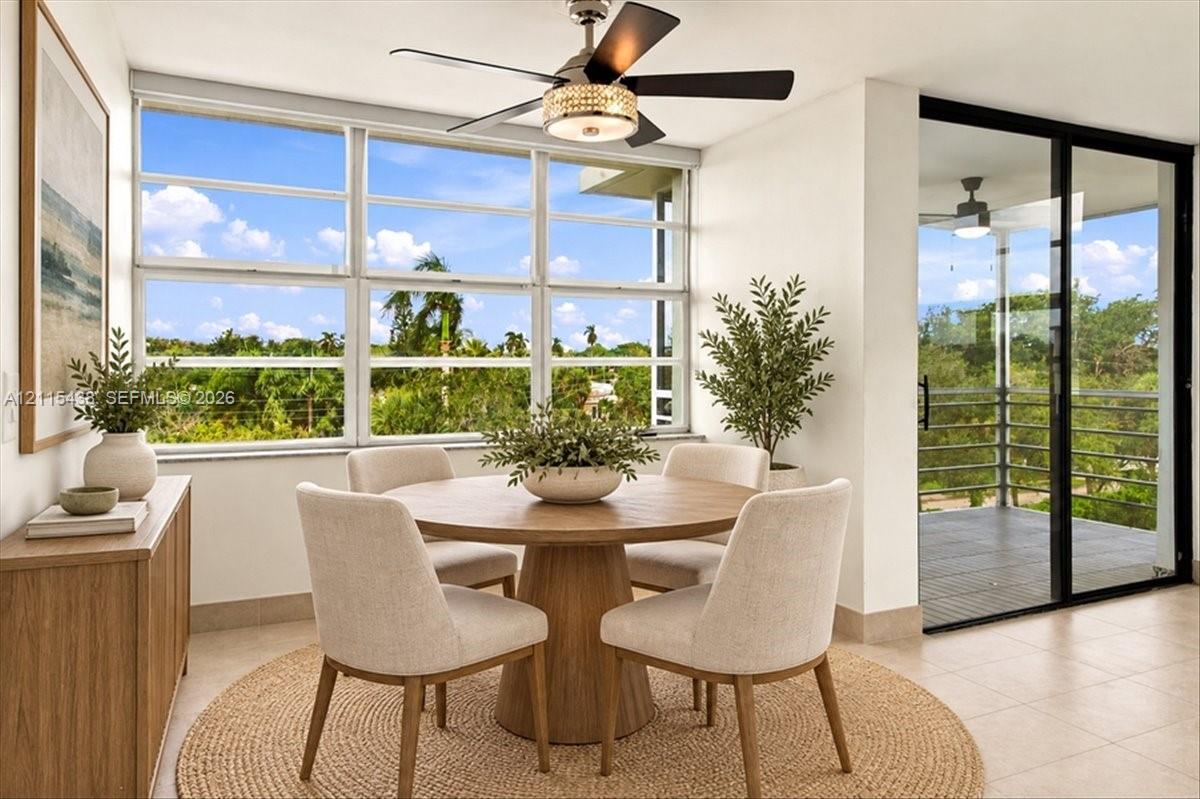 Sun-filled room featuring large, multi-pane windows, neutral tile flooring, a decorative ceiling fan with an integrated light fixture, and a sliding glass door with a metal-railed balcony Sun-filled room featuring large, multi-pane windows, neutral tile flooring, a decorative ceiling fan with an integrated light fixture, and a sliding glass door with a metal-railed balcony