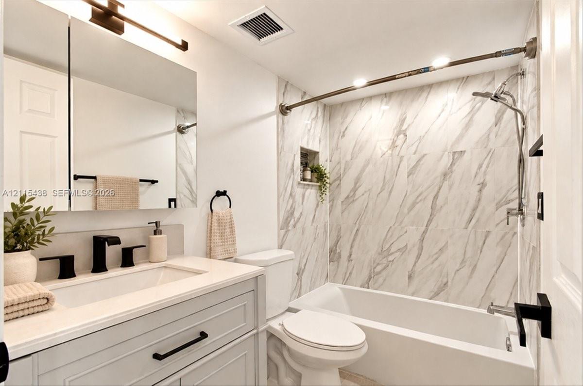 Bathroom featuring a vanity with an integrated sink and light-toned cabinetry, matte black fixtures, a medicine cabinet with mirror, and a tub enclosure with stone-patterned tile surround and a built-in recessed shelf