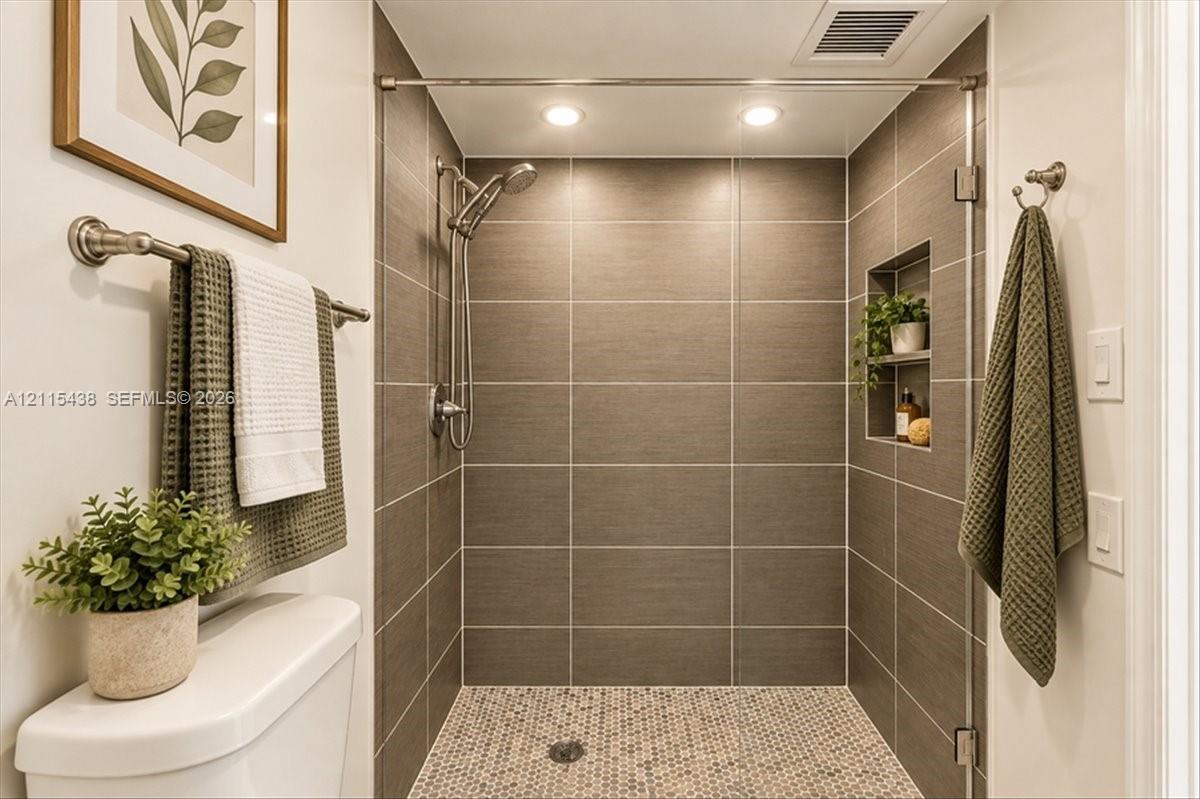 Walk-in shower featuring large-format neutral tile walls, a decorative pebble-style tile floor, a built-in recessed storage niche, and a handheld shower fixture