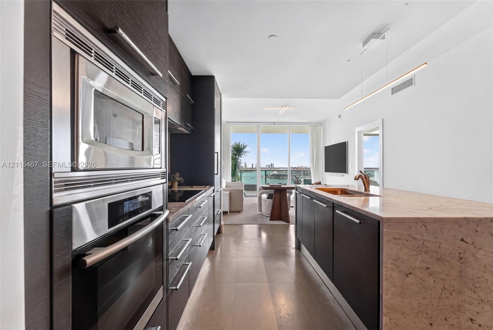 Kitchen featuring dark cabinetry, stone-topped island with integrated sink, stainless steel wall oven and microwave, and linear pendant lighting