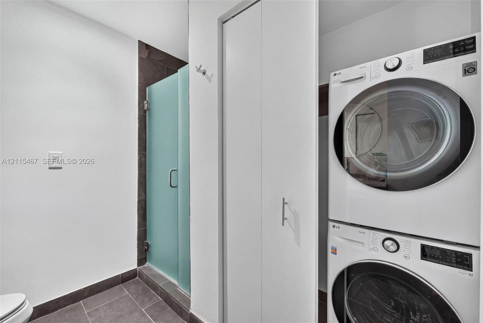 Stacked front-load laundry unit, walk-in shower with a frosted glass door, dark tile flooring, and integrated wall cabinetry