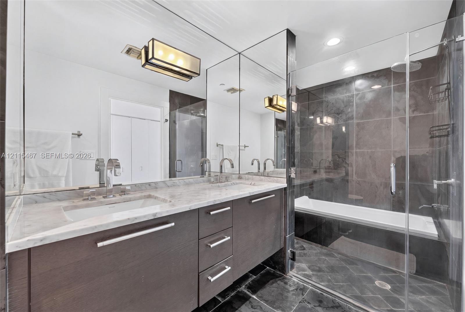 Bathroom featuring a stone-topped vanity with double integrated sinks, dark wood-finish cabinetry, full-width wall mirror, and a glass-enclosed bathtub with large-format tile surround