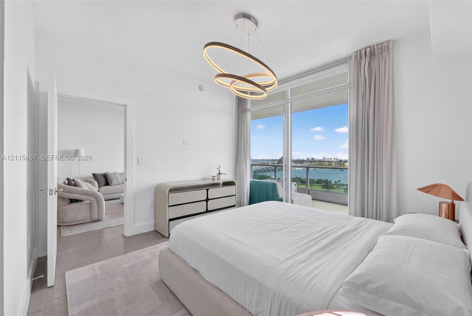 Floor-to-ceiling windows, sculptural multi-ring light fixture, wood-finish flooring, and private balcony with water scenery