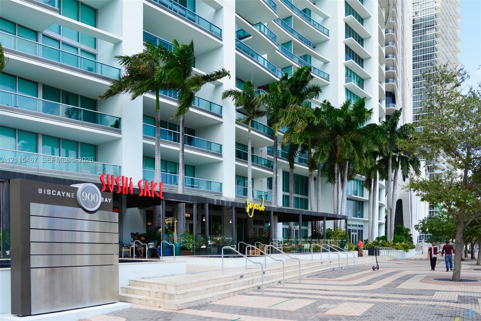 High-rise facade featuring glass-railed balconies and ground-level commercial frontage with stone staircases and palm tree landscaping