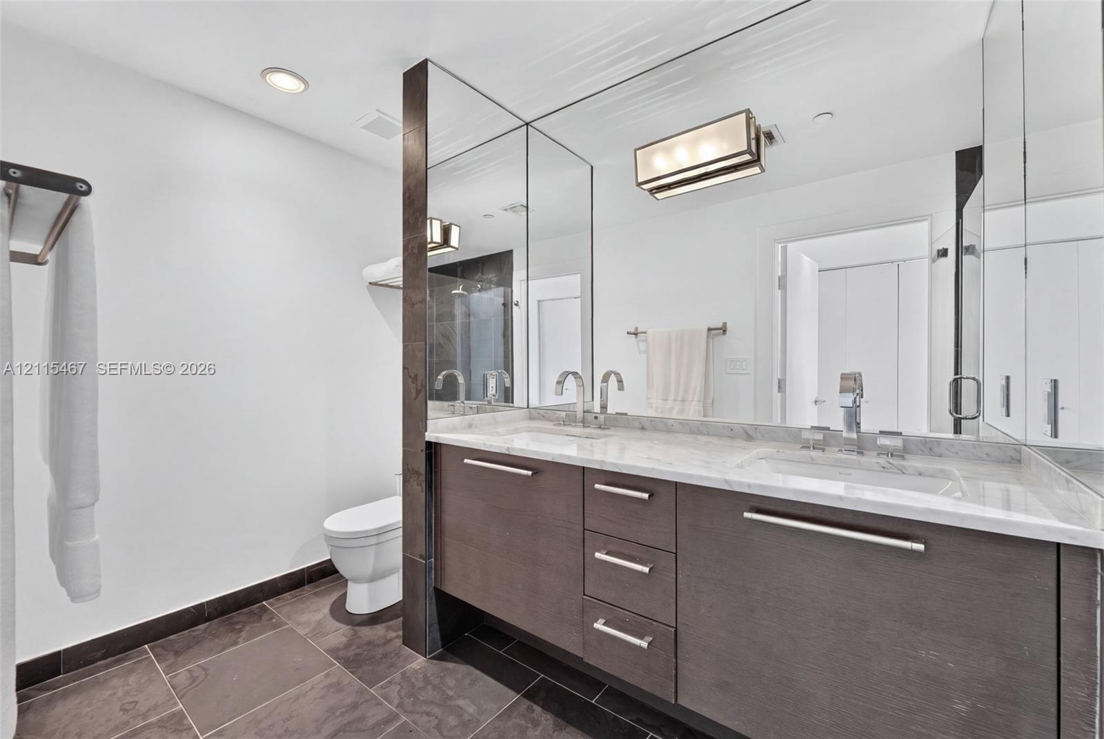 Dual-sink vanity with stone countertop, dark wood-finish cabinetry, full-height wall mirror, and large-format floor tile