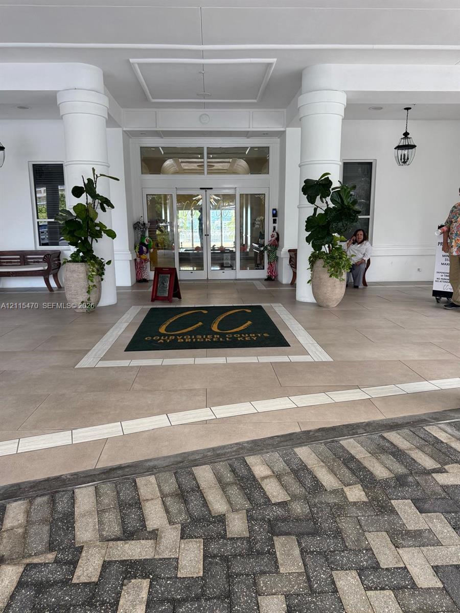 Covered building entrance featuring large structural columns, oversized tile flooring with a central inlay, glass-paned double entry doors, and wall-mounted exterior lanterns