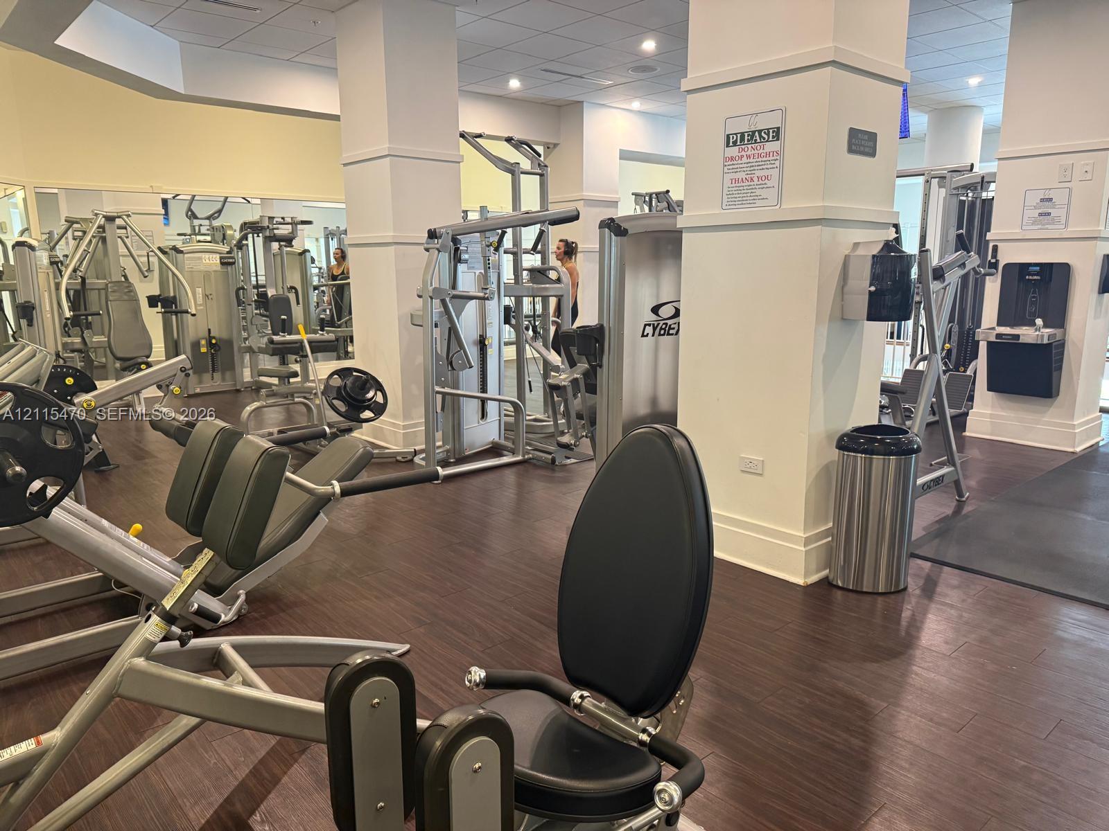 Fitness center featuring wood-finish flooring, structural columns, wall-mounted water dispenser, and multi-station exercise equipment