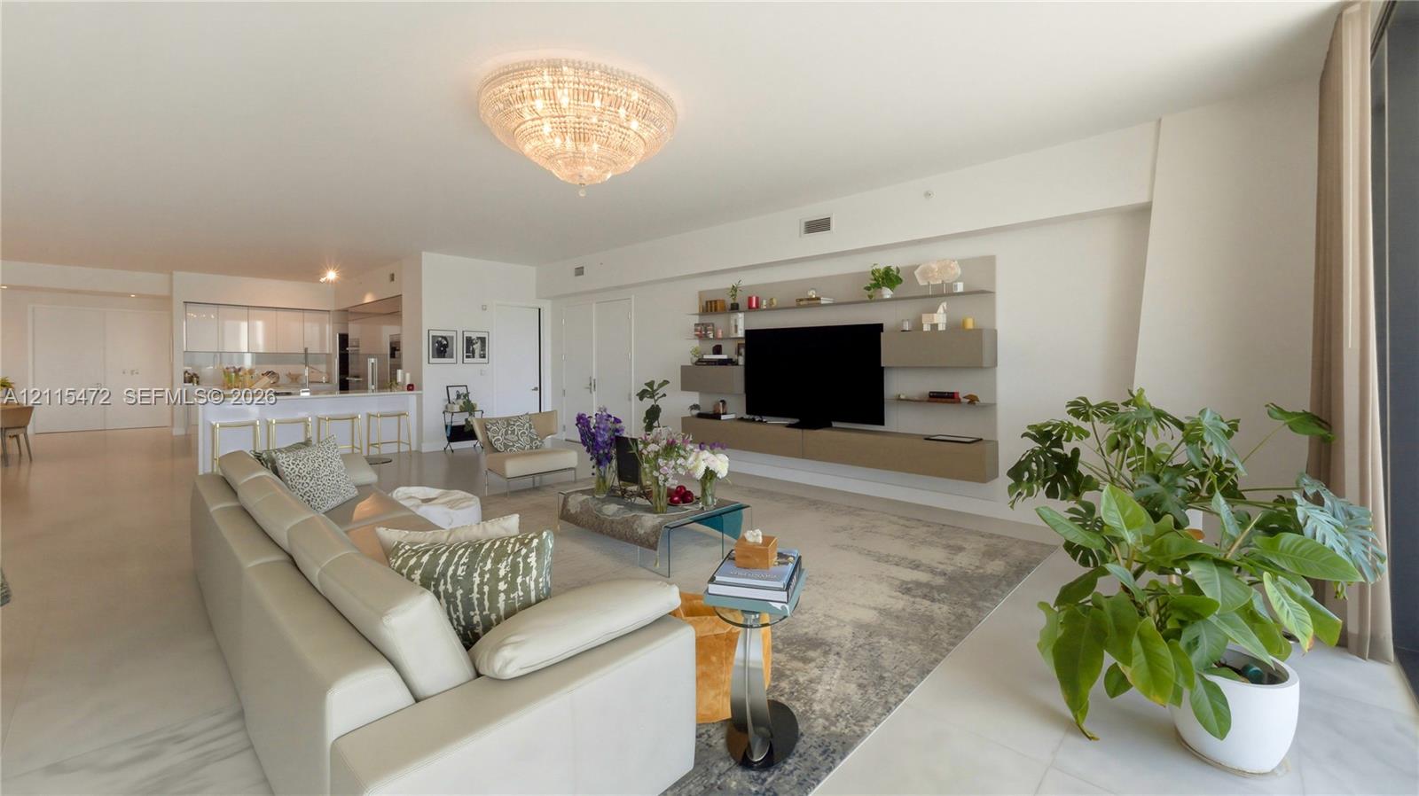 Open-concept living space featuring large-format tile flooring, a statement crystal chandelier, custom wall-mounted floating cabinetry, and a kitchen island with stone-finish countertops