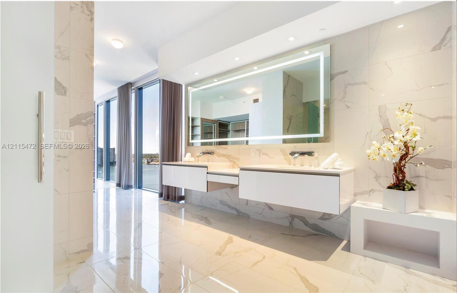Floating dual vanity with integrated sinks, large LED-backlit mirror, floor-to-ceiling stone-look tiling, and expansive floor-to-ceiling windows
