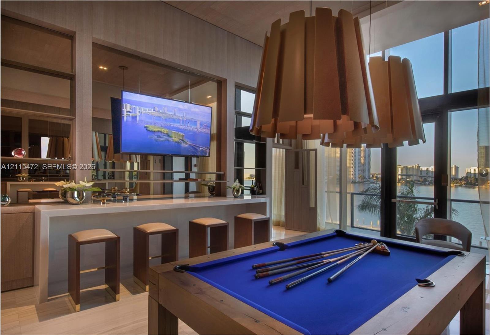 Custom wet bar with integrated cabinetry and stone-surface countertop, floor-to-ceiling glass paneling, oversized geometric pendant lighting, and polished stone flooring
