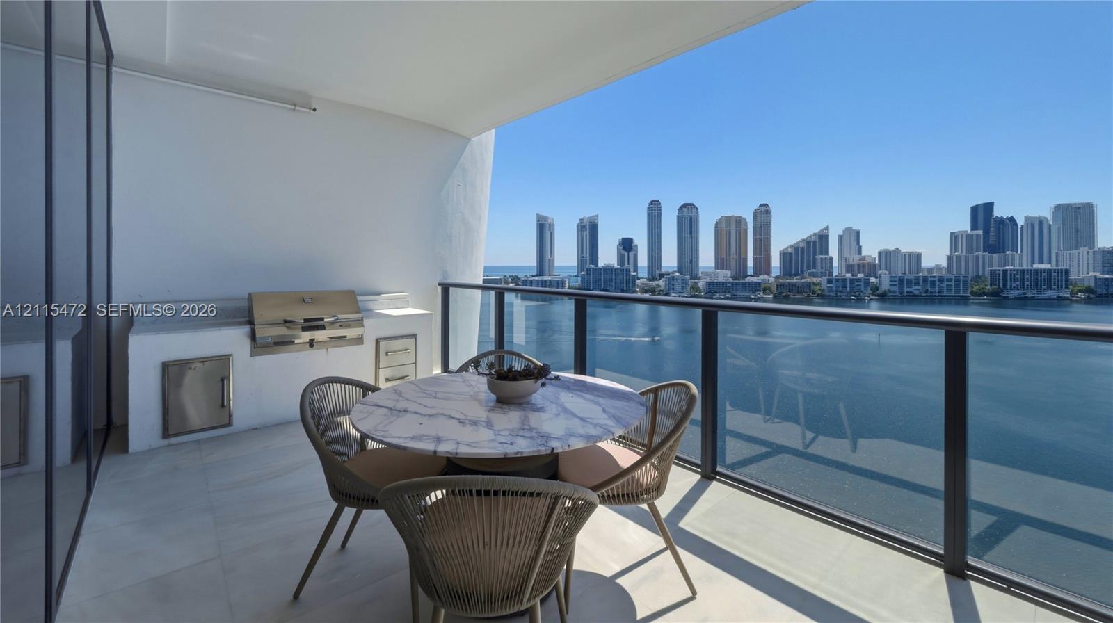Covered balcony featuring a built-in outdoor kitchen with stainless steel appliances, stone countertop, glass railing, and large-format tile flooring