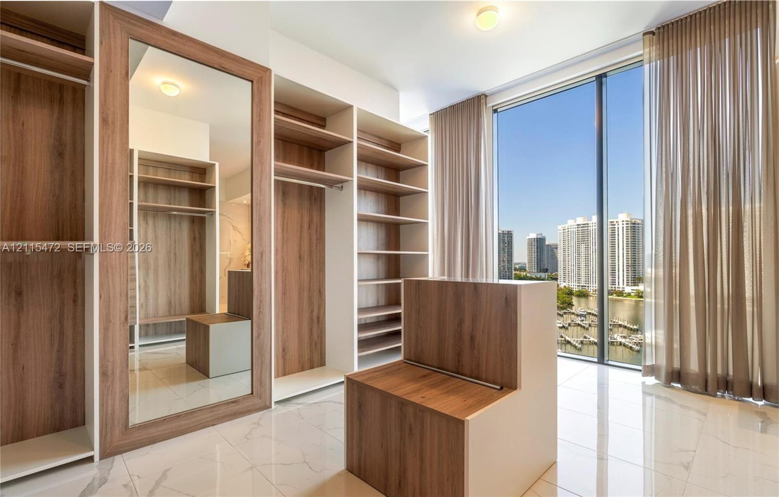 Custom walk-in closet featuring floor-to-ceiling built-in wood-finish shelving, an integrated full-length mirror, polished tile flooring, and expansive window wall with exterior water and high-rise scenery