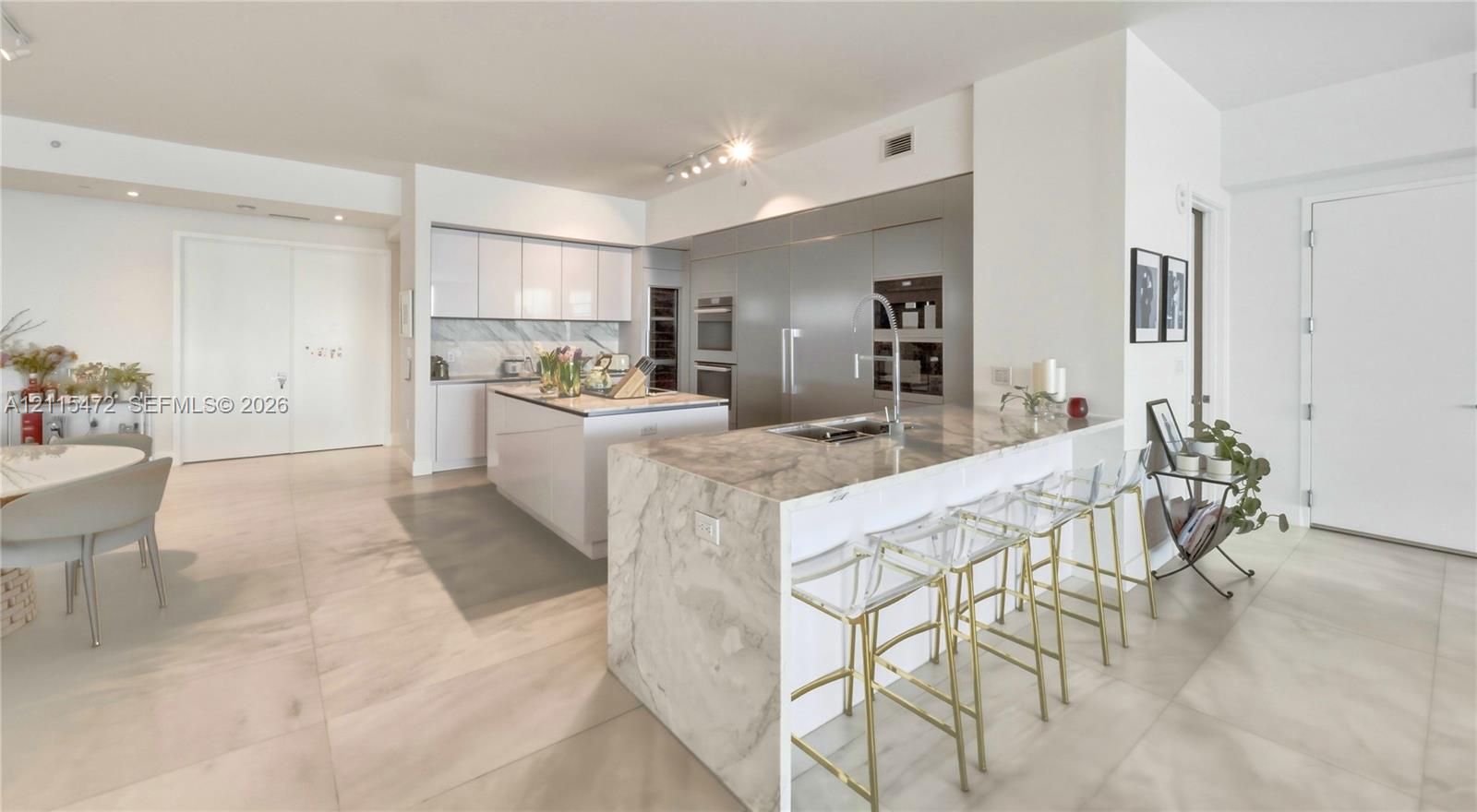 Modern kitchen featuring an expansive waterfall-edge island, stone countertops, integrated stainless steel appliances, flat-panel cabinetry, and large-format tile flooring