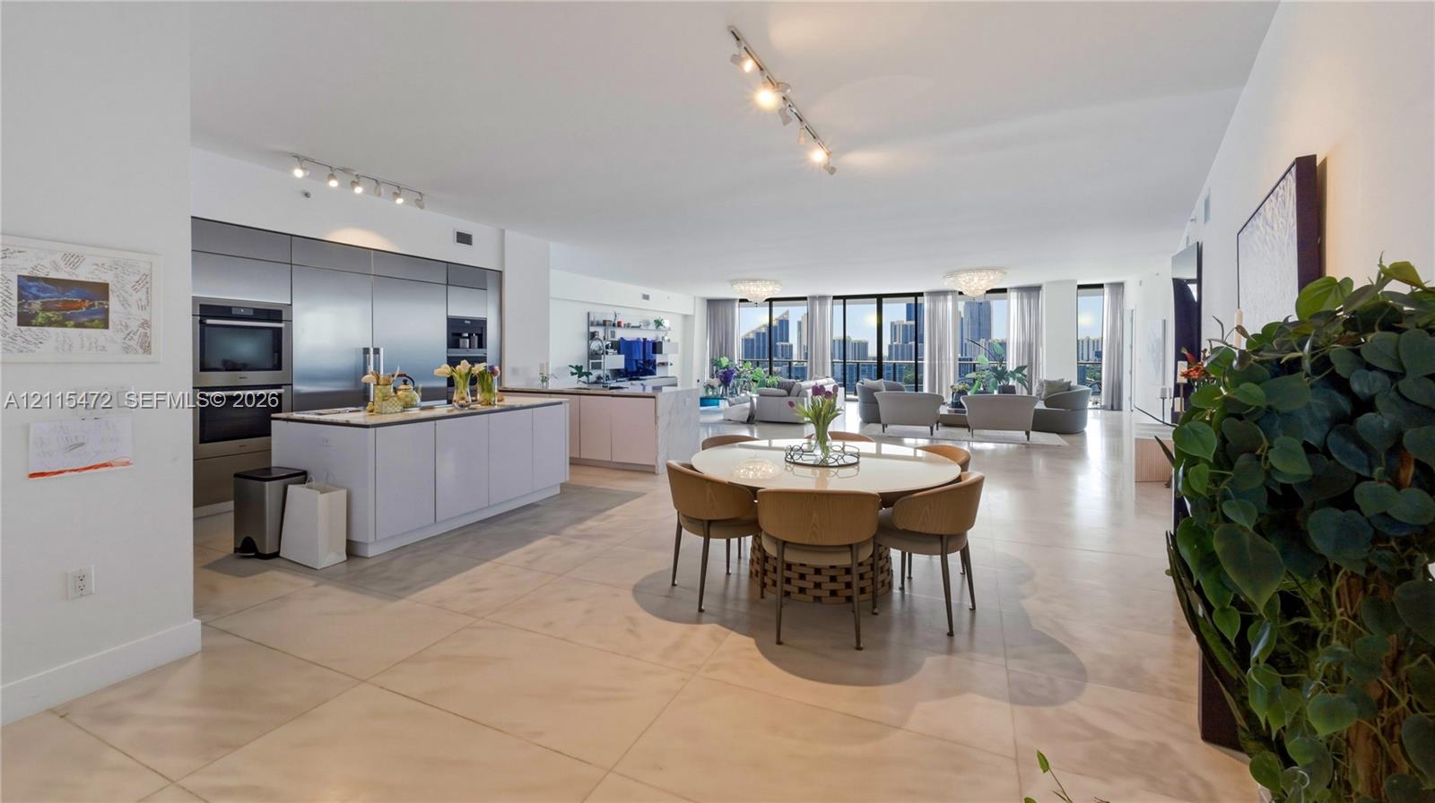 Open-concept kitchen featuring metallic-finish cabinetry, a central island with countertop seating, built-in stainless steel appliances, tile flooring, and floor-to-ceiling windows