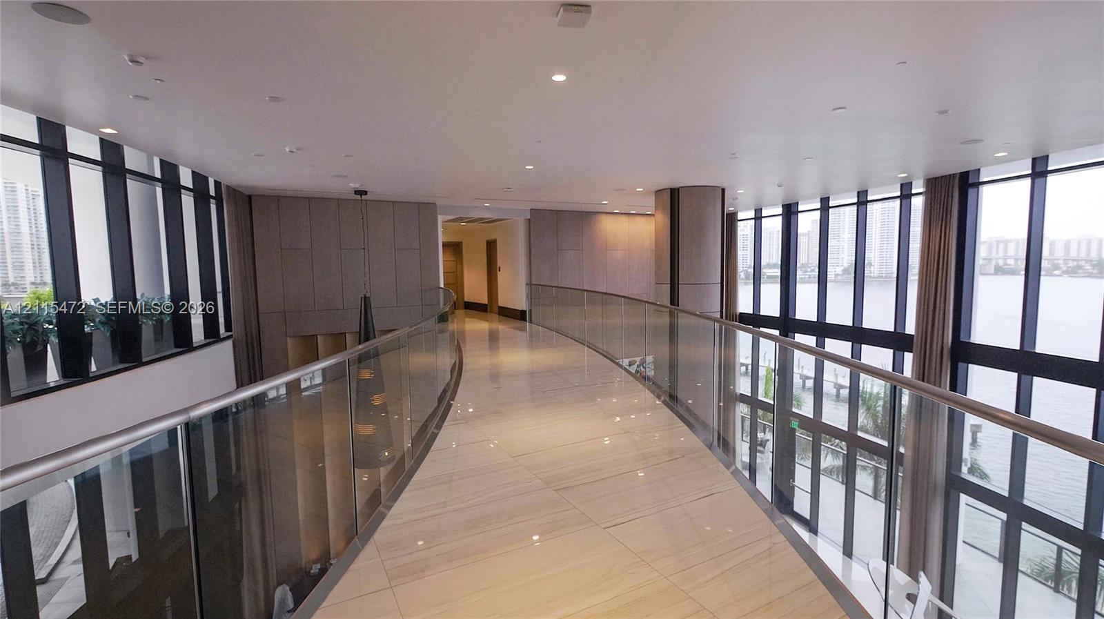 Curved interior bridge featuring floor-to-ceiling windows, polished stone flooring, glass railing, recessed ceiling lighting, and wood-paneled walls