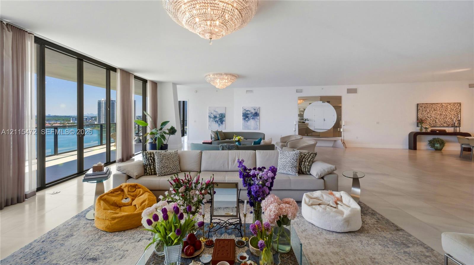 Open-concept living space featuring floor-to-ceiling glass sliding doors, large-format light-toned flooring, two tiered crystal chandeliers, and a circular wall mirror