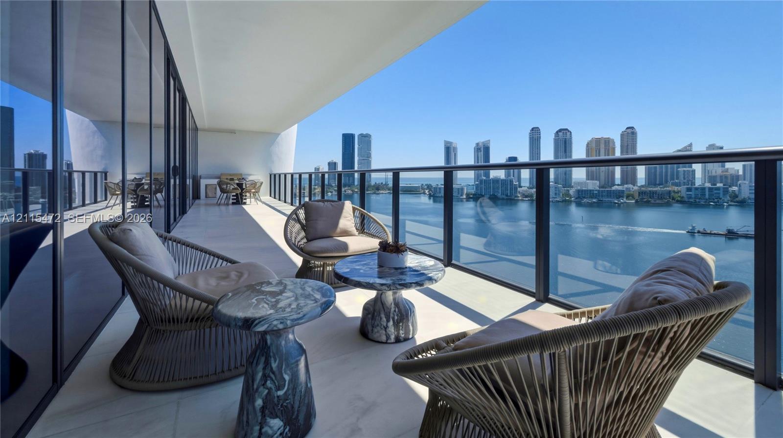 Expansive covered balcony featuring white tile flooring, floor-to-ceiling glass wall, and a full-perimeter metal railing overlooking the water and urban skyline