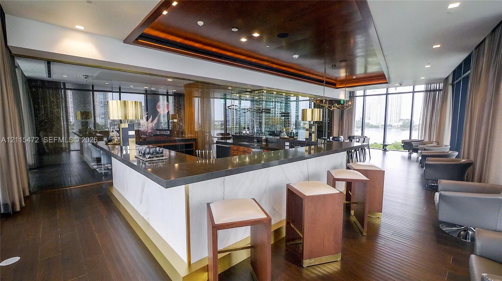 Custom bar featuring a dark stone countertop, stone-paneled base with gold-finish trim, wood-finish flooring, recessed wood-paneled ceiling detail, and floor-to-ceiling glass windows