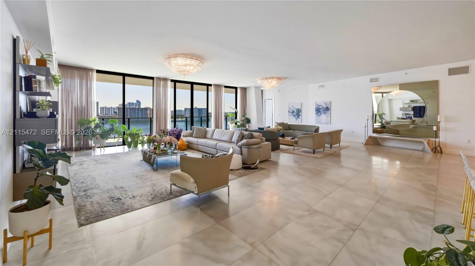 Open-concept living area featuring large-format tile flooring, floor-to-ceiling glass sliding doors, dual crystal flush-mount chandeliers, and a built-in circular wall mirror