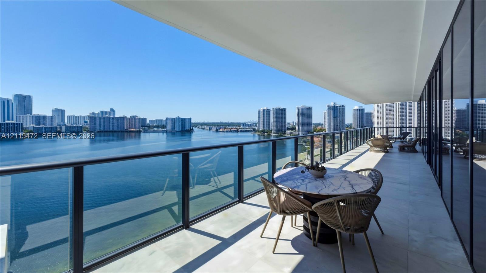 Expansive covered balcony featuring large-format stone tile flooring and a glass-paneled railing, overlooking a coastal waterway and high-rise skyline