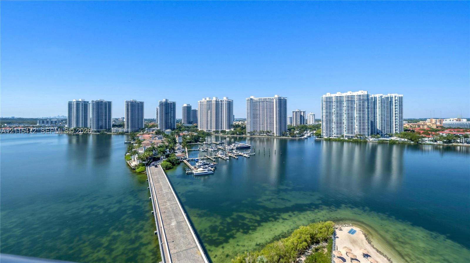 Elevated aerial perspective featuring panoramic water expanses, a private marina with multiple slips, a concrete causeway, and expansive coastal high-rise scenery