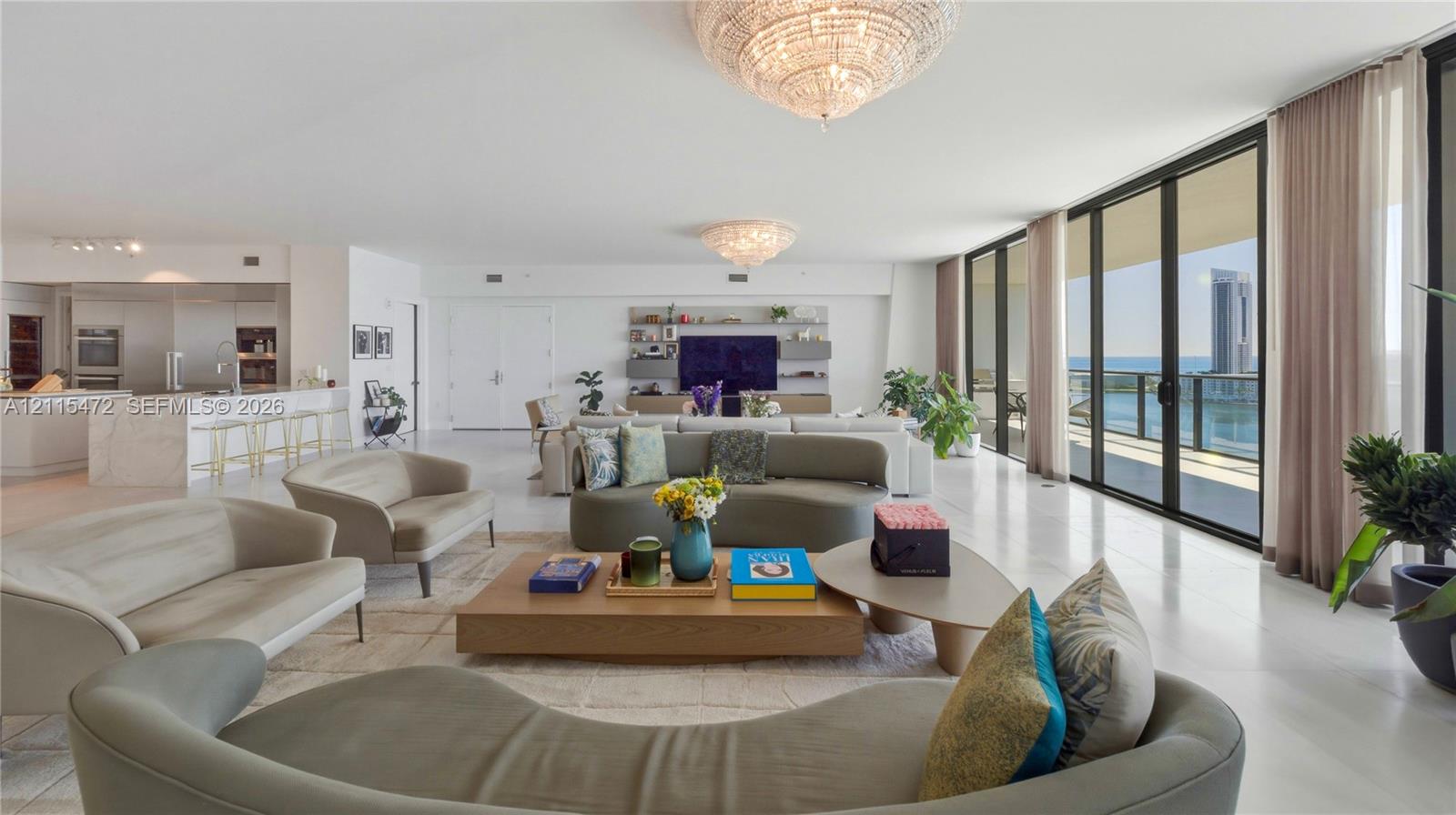 Open-concept living space featuring floor-to-ceiling glass walls, stone flooring, custom built-in wall cabinetry, dual crystal-style chandeliers, and a kitchen island with a stone waterfall edge