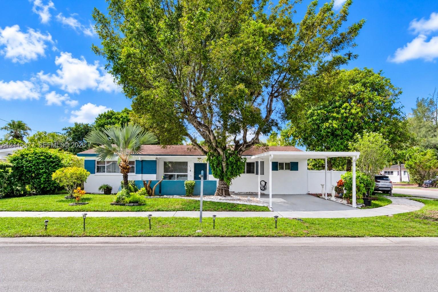 Single-story exterior featuring painted stucco siding, a covered carport, manicured lawn, established shade trees, and privacy fencing