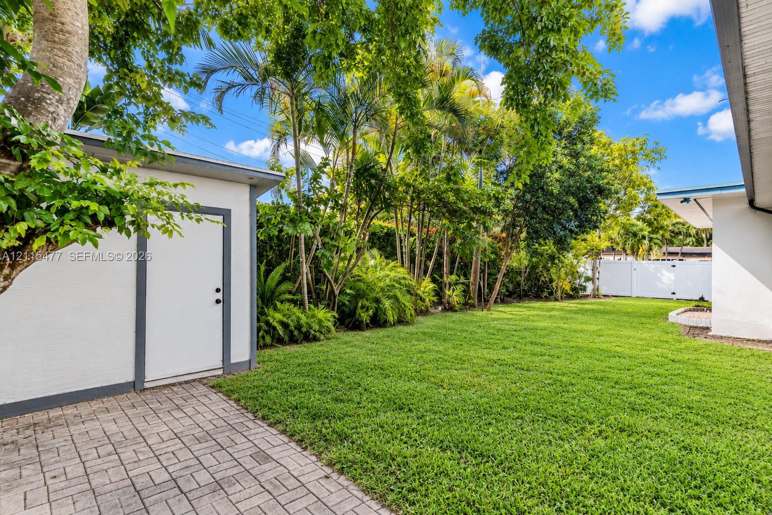 Backyard featuring a paved stone patio, detached storage structure, manicured lawn, and mature perimeter landscaping enclosed by white fencing