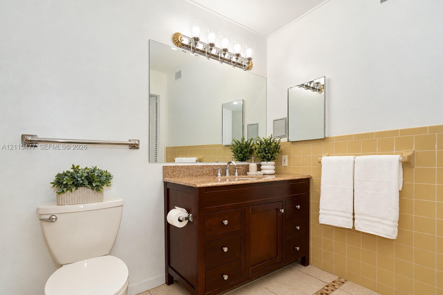 Bathroom featuring a dark wood-finish vanity, stone-surfaced countertop with an integrated basin, wall-to-wall mirror, multi-light fixture, and ceramic tile wall accents