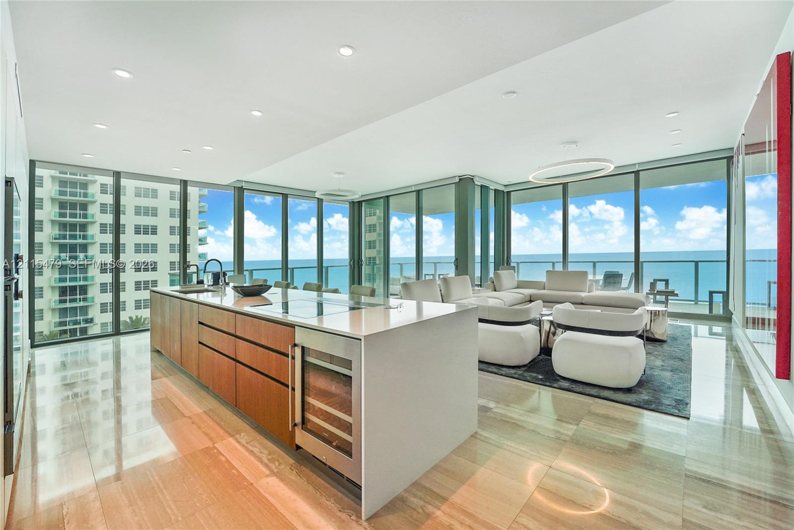 Open-concept kitchen featuring an expansive quartz-topped island, integrated wine refrigeration, warm-tone wood cabinetry, recessed ceiling lighting, and floor-to-ceiling glass panel walls Open-concept kitchen featuring an expansive quartz-topped island, integrated wine refrigeration, warm-tone wood cabinetry, recessed ceiling lighting, and floor-to-ceiling glass panel walls