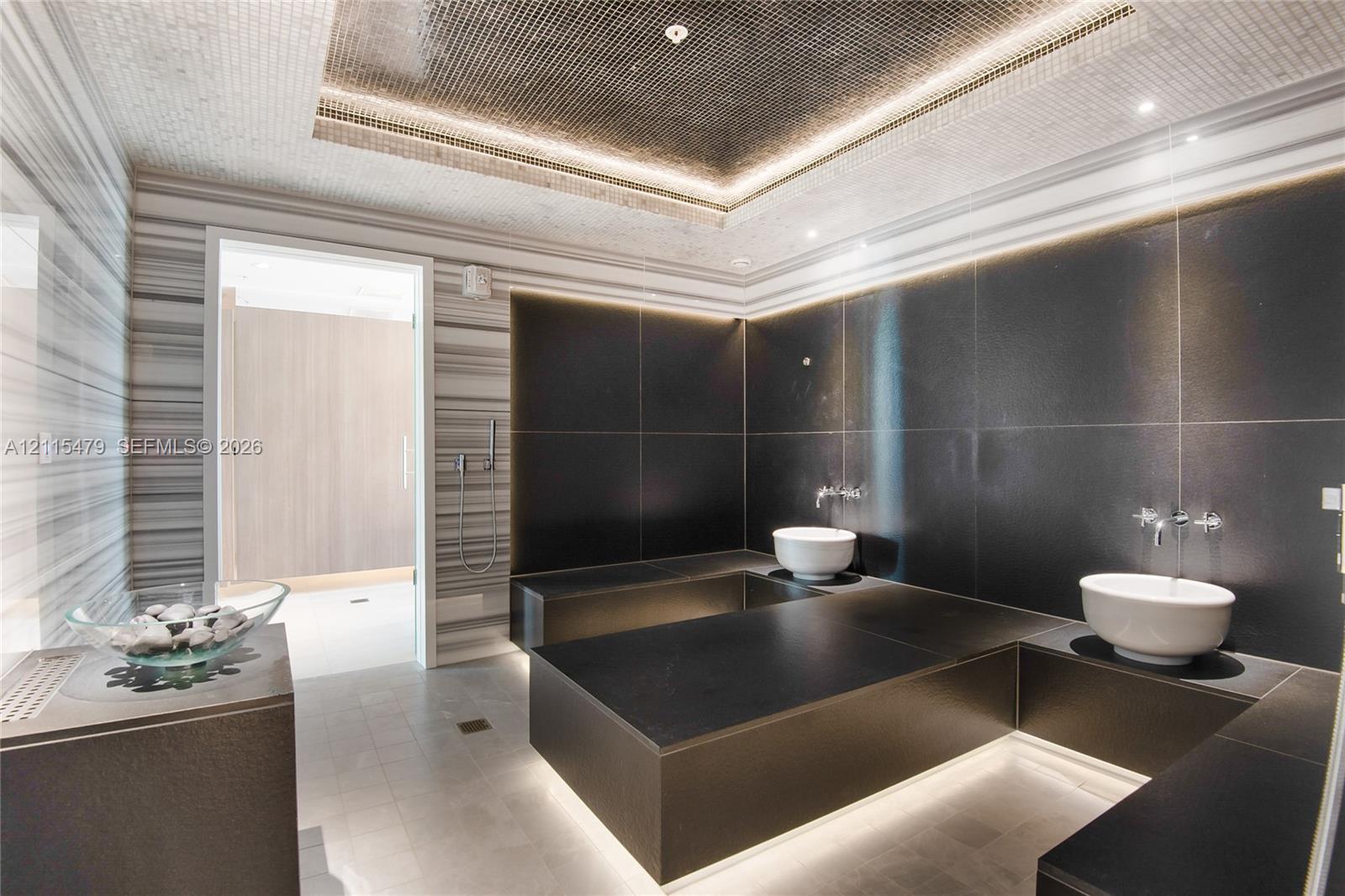 Custom bathroom featuring floor-to-ceiling linear-patterned tile, expansive black-tiled platform surfaces, dual vessel sinks with wall-mounted faucets, and a recessed tray ceiling with integrated perimeter lighting