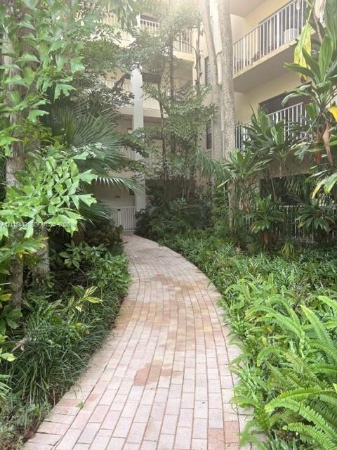 Brick-paved walkway bordered by mature tropical landscaping and established trees, featuring tiered balcony structures with vertical railing systems