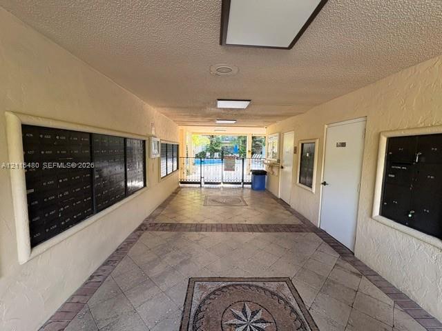 Covered mail station featuring multi-unit metal lockboxes, tile flooring with intricate central inlay, recessed lighting, and a gated entrance