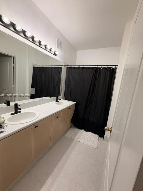 Dual-sink vanity featuring a light-toned wood-finish cabinetry, solid-surface countertop with integrated oval basins, matte black plumbing fixtures, and a large-scale vanity mirror with multi-bulb overhead lighting