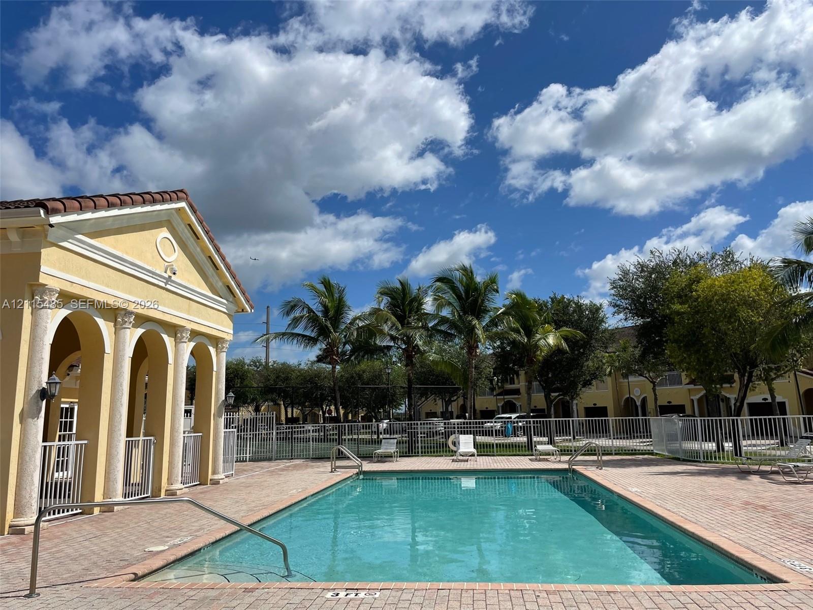 Swimming pool with brick paver decking, enclosed by metal fencing, featuring a columned pavilion with arched entryways and tile roofing