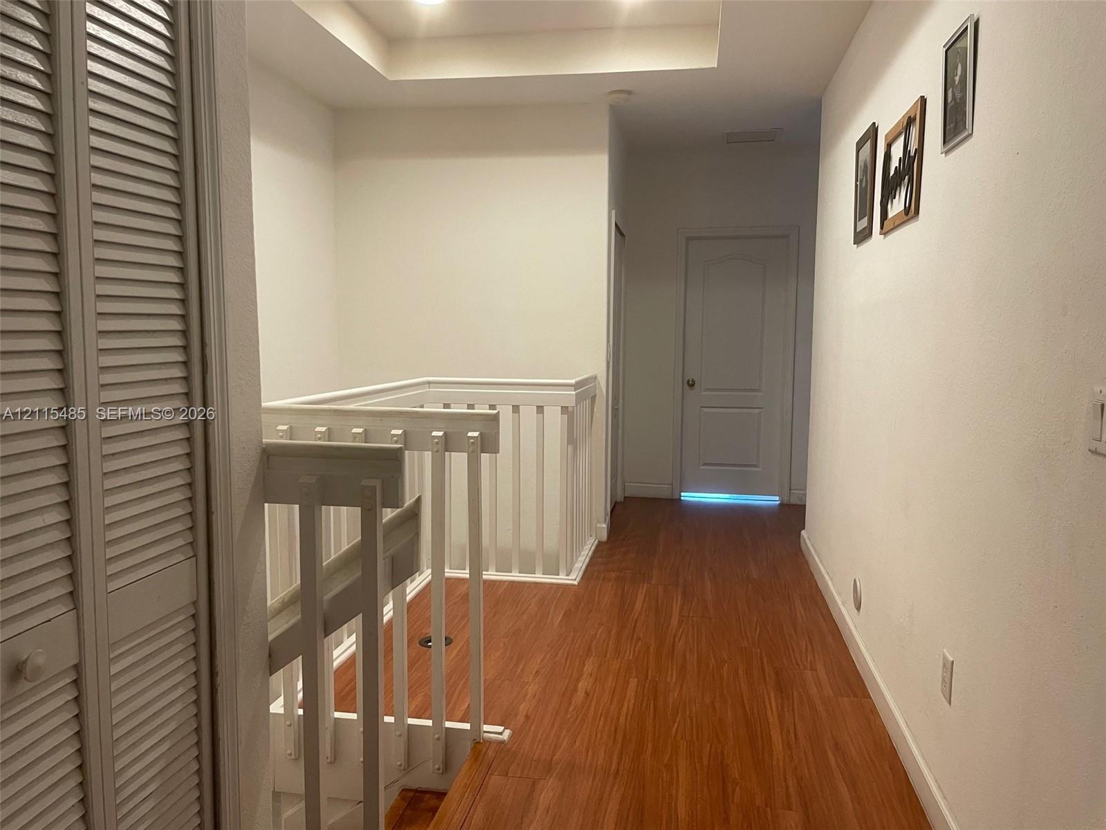 Hallway featuring wood-finish flooring, white louvered-door closet, tray ceiling, and white wooden railing