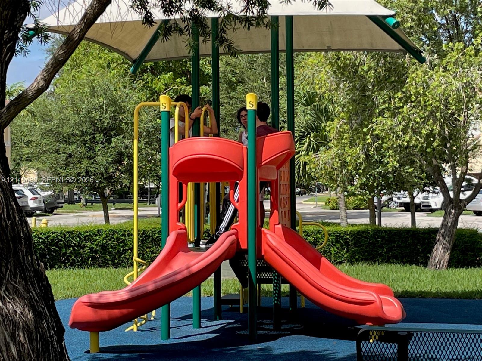 Outdoor recreation area featuring a playground structure with twin slides, a canopy cover, and a rubberized play surface