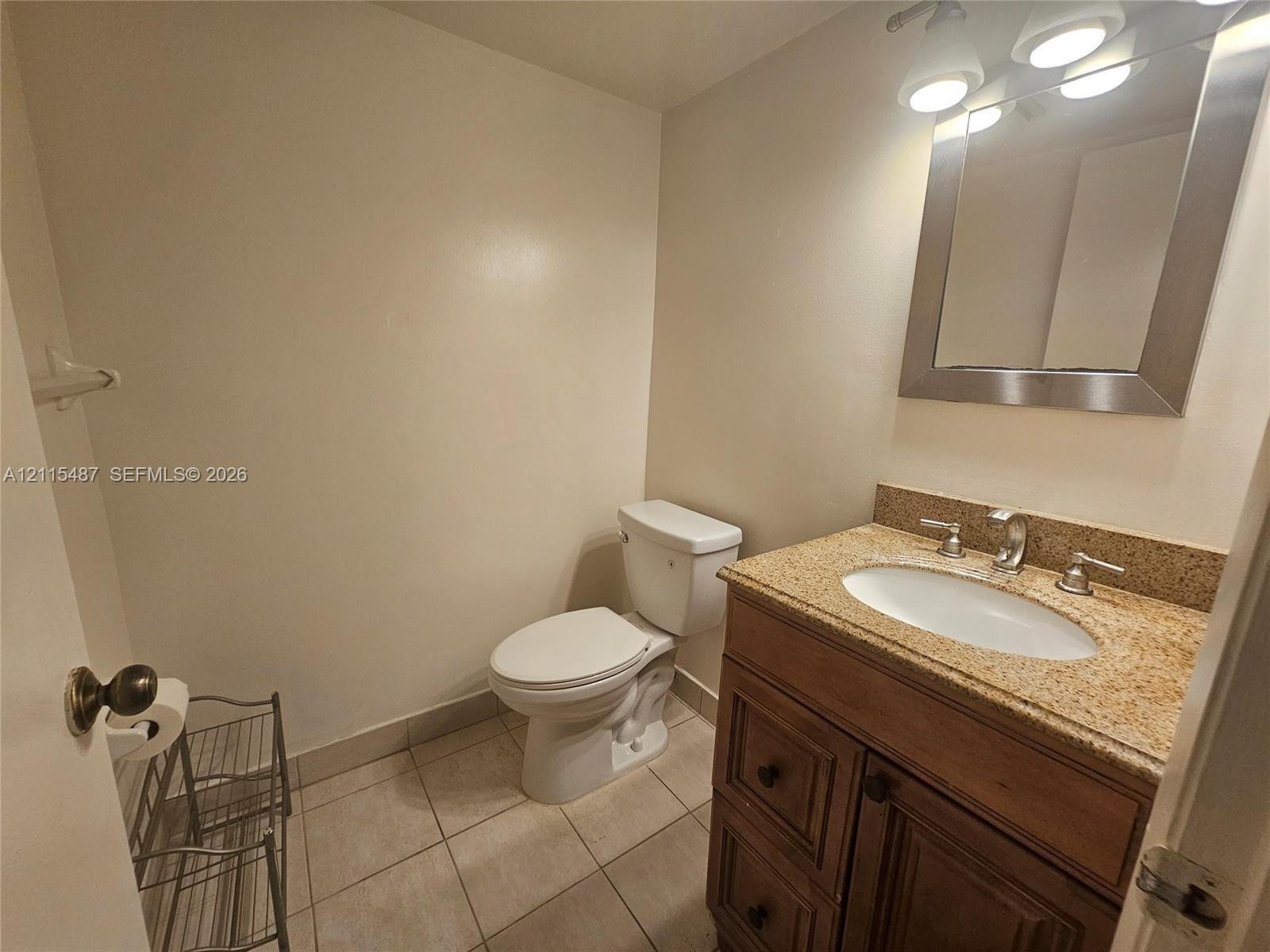 Bathroom featuring a wood-finish vanity with a stone-composite countertop, undermount sink, three-light wall-mounted fixture, and tile flooring