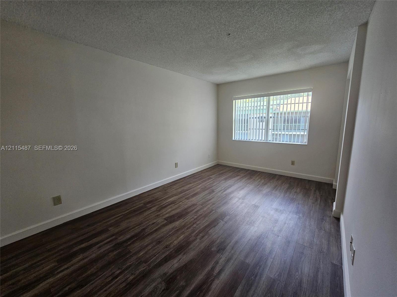 Neutral-toned interior space featuring dark wood-finish flooring, white walls, and a window with horizontal blinds