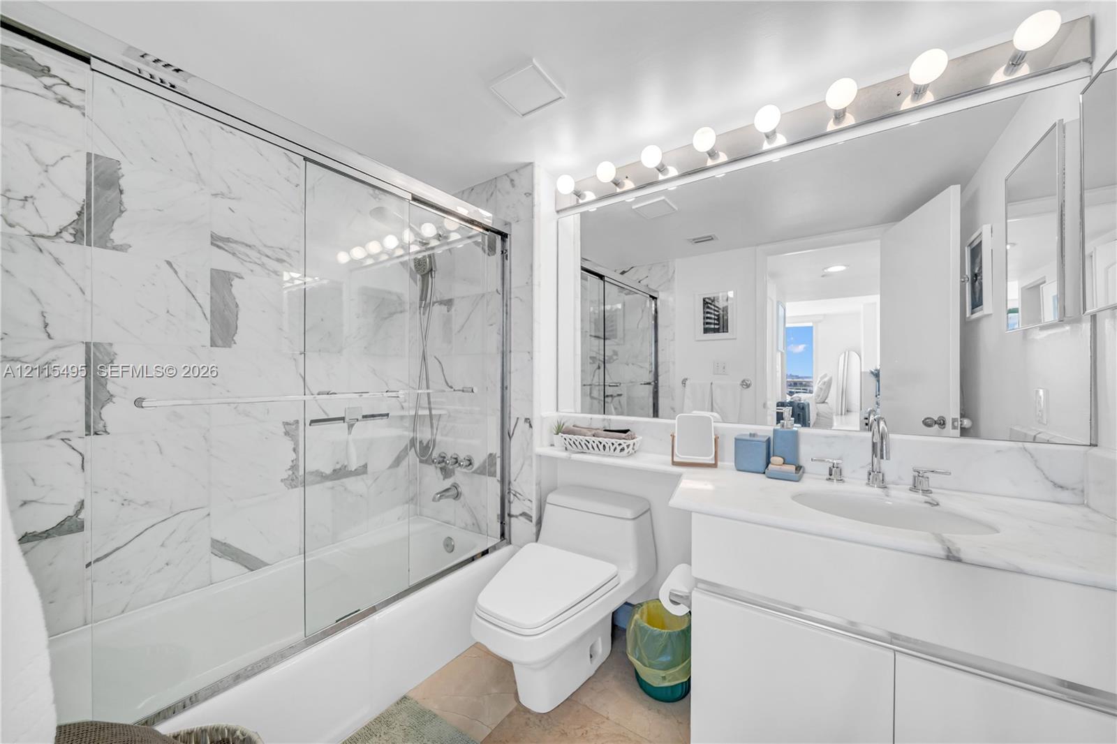 Bathroom featuring a glass-enclosed tub and shower combination with large-format stone-patterned wall tile, white cabinetry with a stone countertop, an integrated oval sink, and a full-width vanity mirror with integrated lighting