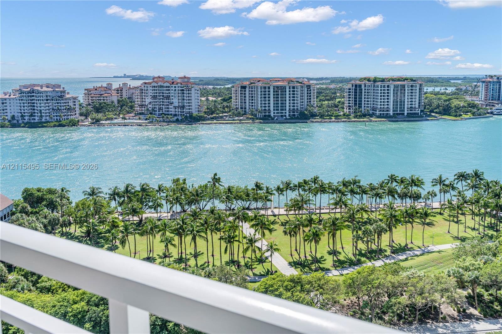 Elevated balcony with a white metal railing, showcasing coastal scenery, waterfront expanse, and a landscaped park with mature palm trees