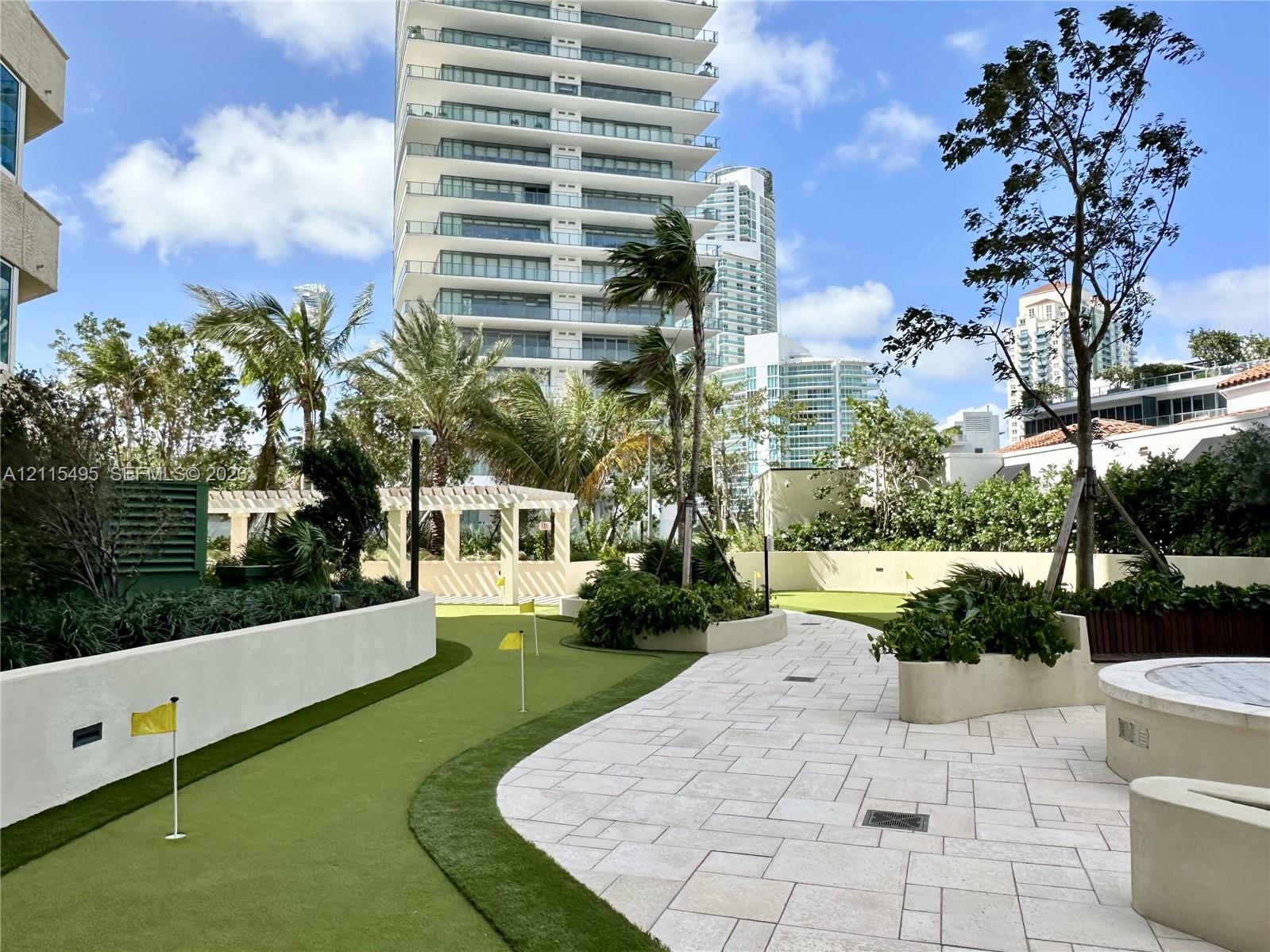 Putting green with flag pins, light-toned stone paved terrace, stucco planter walls, pergola structure, and mature landscaping