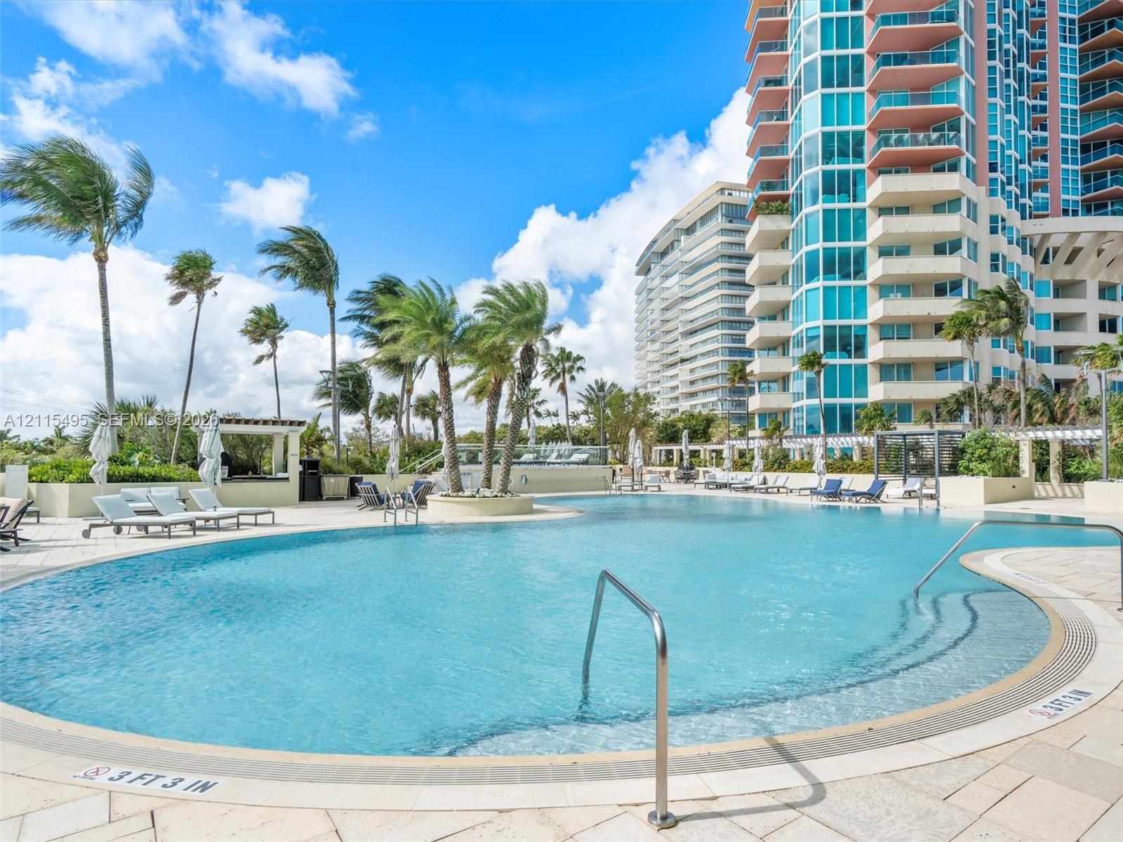 Curved swimming pool featuring a stepped entry, stainless steel handrails, expansive stone-paver decking, and mature palm trees
