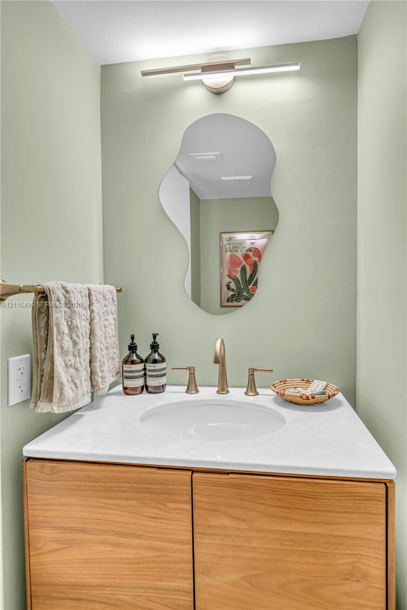 Wood-finish cabinetry topped with a white solid-surface vanity, integrated undermount sink, brass-finish widespread faucet, organic-shaped wall mirror, and contemporary linear light fixture
