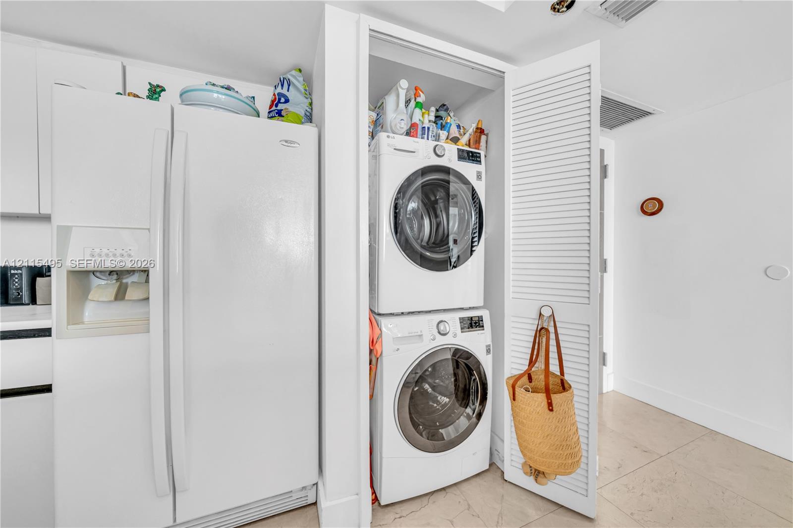 Stacked laundry unit housed within a closet featuring white louvered doors, positioned beside a full-size refrigerator with an external water and ice dispenser, set against stone-finish flooring