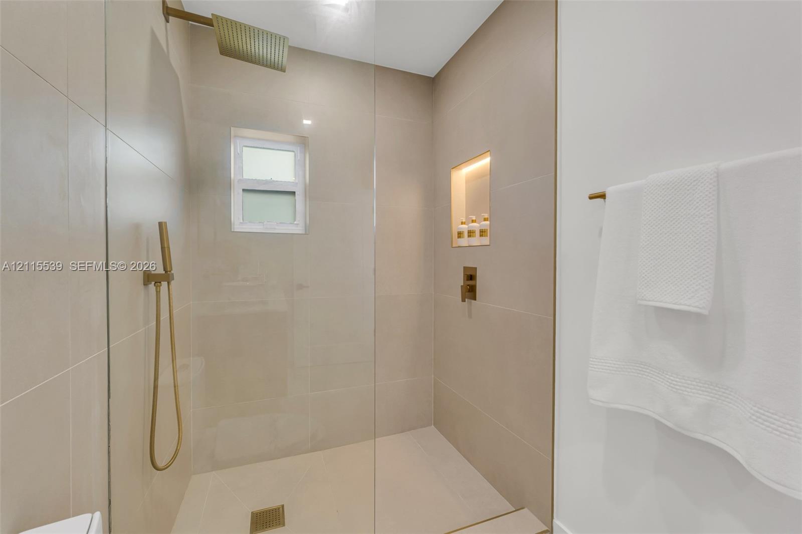 Walk-in shower featuring large-format wall tile, a frameless glass enclosure, a rainfall showerhead, a handheld sprayer in a metallic finish, and a recessed tiled niche with integrated lighting