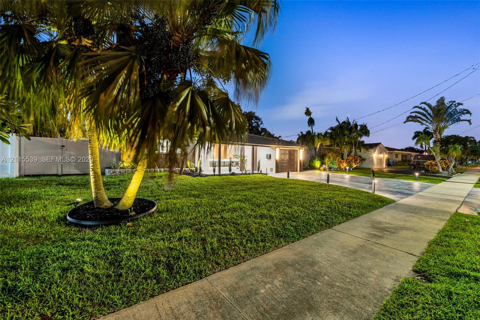 Exterior facade featuring a concrete driveway, manicured lawn, mature palm trees, white vinyl privacy fencing, and exterior landscape lighting